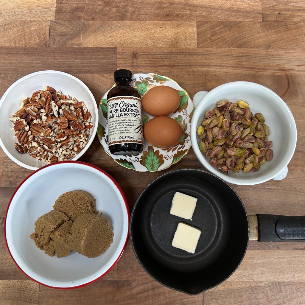 Little plates filled with pecans, eggs, pistachios, and brown sugar, alongside a bottle of vanilla and a small pan with two pats of butter.